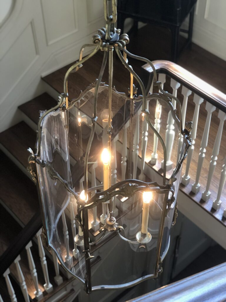 A brass and glass lantern-style chandelier with lit bulbs hangs over a wooden staircase with white railings.