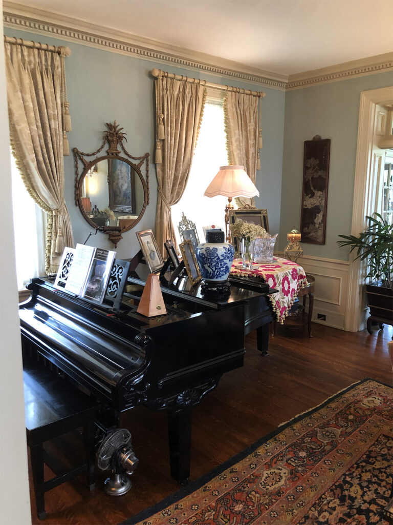 A grand piano with sheet music and framed photos sits in a traditional living room with ornate decor and wooden floors.